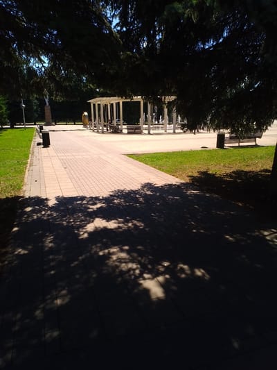 The image depicts a shaded paved walkway in a park in Otradny, Russia, with dappled sunlight filtering through overhead trees. In the background, a white pergola with benches is visible, and further beyond, a series of white fountains are in operation. A statue on a pedestal stands to the left, and dense green trees line the perimeter of the park. The walkway is constructed of light brown paving stones, and the surrounding area is covered in green grass. Two dark-colored trash receptacles are positioned along the edge of the walkway.
