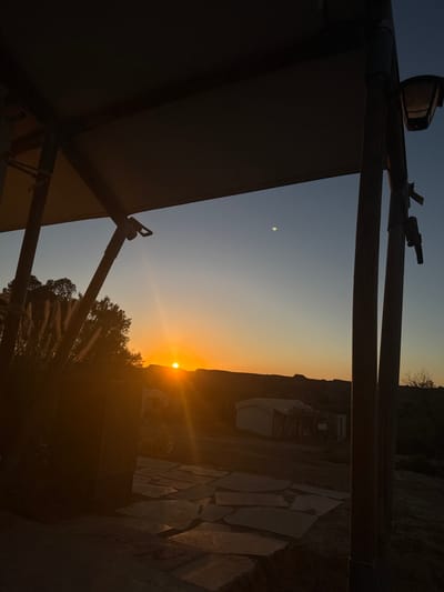 The image depicts an outdoor landscape scene during either sunrise or sunset in the Merhavim Regional Council, Israel. The foreground is dominated by the dark silhouette of a structural overhang, likely part of an awning or canopy, supported by two vertical poles visible on the left and right. A light fixture is attached to the rightmost pole. Below the overhang, on the ground, an irregular stone slab pathway or patio is visible. To the left, a cluster of tall, feathery grasses or similar vegetation stands, backlit by the sun. The midground features an arid, undulating terrain with sparse vegetation and at least two small, light-colored structures, possibly tents or cabins. In the background, low-lying hills or mountains are silhouetted against the horizon. The sun is a prominent, intense orange orb positioned just above these distant hills, casting strong rays across the scene. The sky transitions from a brilliant orange and yellow at the horizon to a clear, deep blue at the top. A distinct, small, bright off-white celestial body, possibly the Moon or a planet, is visible in the upper-right portion of the blue sky. There are no people present, and no active interactions or specific actions are depicted.