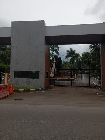 The image shows a gated entrance to a property in Kecamatan Kutawaringin, Indonesia. The gate is made of black metal bars and is closed. There is a guard booth on the left side of the gate. The booth is made of concrete and has a small window.  The booth is topped with a gray roof.  There is a red and white traffic cone on the left side of the guard booth. The gate is under a large, white, concrete archway with a brown wood accent on the top. The gate is flanked by two concrete pillars. The gate is covered in red and black paint. Behind the gate, a paved road leads to a small statue of a man in the middle distance. The statue is located on the left side of the road. The man is wearing a gold-colored outfit. On the right side of the road, there is a green area with trees. The sky is gray and cloudy. The road leading to the gate is paved and appears to be gray asphalt.