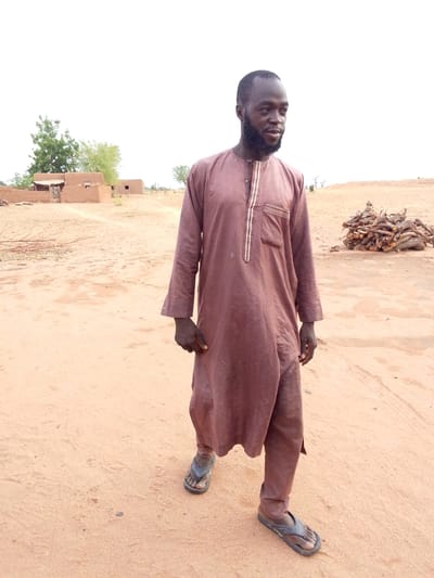 A man stands in a sandy, arid environment in Mulke Barikin Sayejo, Nigeria. He wears a long, brown robe-like garment and simple sandals. Behind him are low, mud-brick buildings and a pile of cut wood. The sky is overcast.