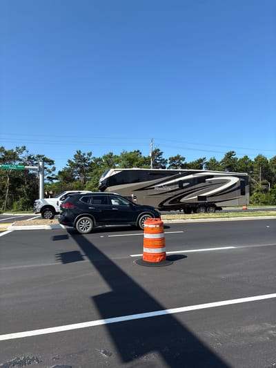 On a clear, sunny day in Panama City Beach, Florida, a black SUV is parked on the right side of a road, facing left. Behind the SUV, a large recreational vehicle (RV) is parked. The RV is gray and black with a decorative swirl pattern and the word "Montana" on its side. To the left of the SUV and RV, a white pickup truck is parked. A traffic signal pole is visible to the left of the vehicles, with a green street sign that reads "Panama City Beach Parkway." In the foreground, an orange traffic barrel with white stripes sits on the asphalt road. The road is marked with white lines. Trees and power lines are visible in the background.