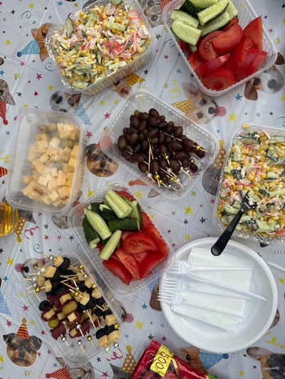 The image displays an outdoor picnic table set with various food items and party decorations. The table is covered with a white plastic tablecloth featuring cartoon images of dogs and cats wearing party hats, as well as colorful stars and confetti.

Arranged on the table are several clear plastic containers holding food. In the top left is a container filled with a salad containing crab sticks, corn, and chopped cucumbers in a creamy dressing. To its right is a container of sliced tomatoes and cucumbers. Below the crab salad, a container holds cubed yellow cheese and green olives. Adjacent to the cheese is a container of dark purple grapes, some of which are skewered with decorative toothpicks. In the bottom left is a clear container filled with skewers of cubed cheese, sliced salami, black olives, and green olives, also adorned with decorative beads. To the right of the grapes is another container of crab stick salad, similar to the one in the top left.

A white plastic plate is positioned in the lower right quadrant of the image, holding a stack of white paper napkins and three white plastic forks. A black plastic fork rests on the crab salad container. A glass with a golden liquid, possibly juice or beer, is partially visible on the left side of the image. A red and yellow bag with Cyrillic text and the number "500" is also present in the bottom right corner.

The overall scene suggests a casual outdoor gathering or party. The specific location in the city of Valok, Russia, cannot be determined from the visual information provided in the image.