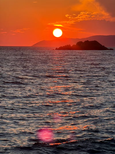A vibrant sunset illuminates the Adriatic Sea off the coast of Mirište, Montenegro. The sun, a brilliant white orb, hangs low in the sky, casting a warm, orange glow that suffuses the clouds and the water. A small, rocky island, silhouetted against the fiery sky, sits in the middle distance. The sea is a deep, dark blue, its surface rippled with gentle waves that catch the reflected light of the sunset, creating shimmering streaks of orange and pink. The overall scene is one of tranquil beauty, with the natural elements of sun, sea, and land converging to create a breathtaking vista.