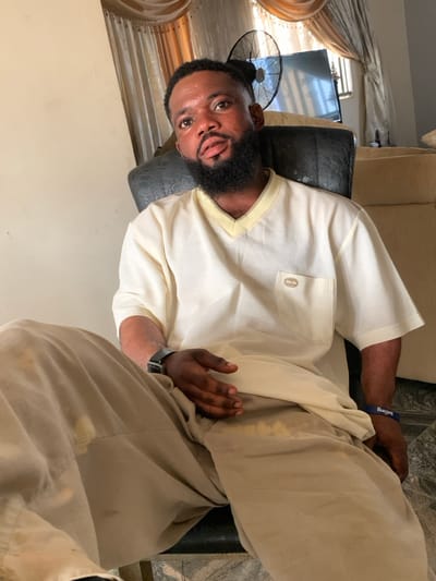 The scene is situated within Eika-Adagu New Layout, Nigeria. An adult male is seated indoors, positioned centrally and facing the viewer directly. He has a dark, well-groomed beard and mustache, with short, dark hair. He wears a light cream-colored, short-sleeved V-neck top featuring a small pocket with a light brown label on the left chest. His lower garment consists of light brown or khaki trousers, displaying discolouration or stains on the left thigh and groin area. A dark-colored smartwatch with a black strap is on his left wrist, and a blue wristband with partial white text is on his right wrist. His left hand rests open on his left thigh. The man is seated in a dark-colored chair or sofa. In the background, a dark standing fan with an open cage is visible to the right of the man's head, partially obscuring a wall-mounted flat-screen television. Cream-colored curtains with decorative valances are present in the background. A light-colored wall is to the left of the subject, and tiled flooring, possibly marble or polished stone, is visible at the bottom of the frame. A portion of another light-colored sofa is discernible in the far right background.
