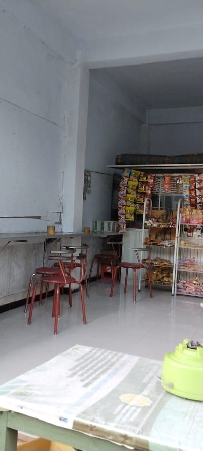 The image depicts the interior of a small shop in Mataram, Indonesia, likely a convenience store or a small warung. The shop appears to be open to the street or an adjacent space, as indicated by the open doorway on the right side of the frame. Inside, the walls are painted a light, neutral color, possibly white or pale gray, and show signs of age and wear, with some scuff marks and minor damage visible.
The flooring is tiled with light-colored square tiles. In the foreground, a table covered with a white sheet or tablecloth, printed with text and images, is partially visible. A bright green plastic object, possibly a fan or a similar small appliance, rests on the covered table.
The main area of the shop is dominated by shelves and displays of packaged goods. On the right side, a tall metal shelving unit is stocked with various food items, including bags of snacks, drinks, and other packaged products. The items are arranged in rows and appear to be commonly found in Indonesian convenience stores.
In the center and left of the image, there are a few simple red metal chairs with round seats and thin metal legs, arranged around small tables or counter spaces. These chairs suggest a seating area for customers. Some of these chairs are stacked or pushed together.
Hanging from the wall or the shelves are more packaged goods, including what appear to be small bags of chips or candy, arranged vertically. There are also some white plastic baskets on a shelf or counter.
The lighting in the shop seems to be a mix of natural light coming from the open space and possibly some internal lighting, creating a somewhat dim but functional atmosphere. The overall impression is of a typical, no-frills local shop serving everyday needs.