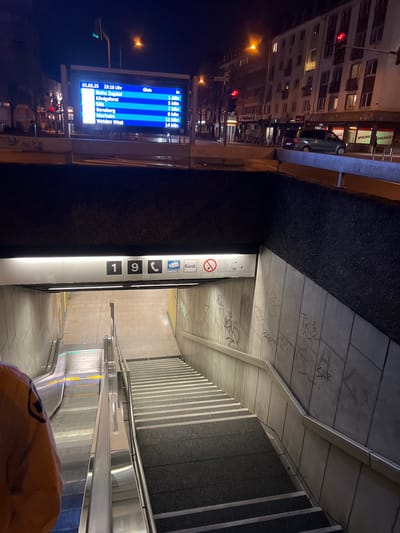 The image shows an entrance to an underground station in Köln, Germany, at coordinates 50.939, 7.009. There is a sign above the entrance displaying the number "19". The entrance is located under a street-level walkway, and is also visible from the street. There are stairs and an escalator leading down into the station. The station entrance is partially obscured by a person wearing an orange jacket. Graffiti is visible on the walls of the passageway. There is a lit sign above the entrance with "Kiosk" and a phone icon.  There is a digital sign displaying train departure information, showing the time and destinations.  The scene is lit by streetlights and interior station lights.  The environment is urban.