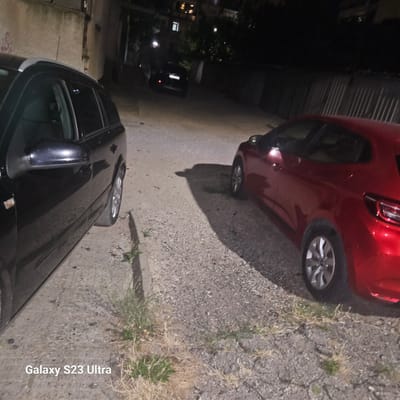 In the city of Haskovo, Bulgaria, an outdoor scene at night captures two cars parked on a gravelly surface with some dry grass and weeds. On the left, a black station wagon is parked with its side visible, featuring a roof rack and a side mirror. To its right and slightly further back, a red hatchback is parked, showing its rear passenger side and wheel. In the background, a dark, narrow alleyway is visible, with a building on the left and a corrugated metal fence on the right. Faint lights from apartment windows are visible on a building in the far distance. The ground is uneven and composed of gravel and dirt. A watermark at the bottom left of the image reads "Galaxy S23 Ultra."