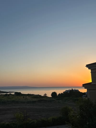 This is a vertical, eye-level shot of a coastal sunset scene in Chernomorets, Bulgaria. The sky transitions from a clear, pale blue at the top to a vibrant orange glow near the horizon. The sea is calm and reflects the sunset colors. In the foreground, there is a construction site with dark, barren soil and several fences. Behind the construction site, there is a patch of green grass and some scattered trees. On the right side of the image, a portion of a building's exterior is visible, featuring a beige facade and a reddish-brown tiled roof. In the distance, along the coastline, there are some low-lying hills or landmasses. The overall mood is serene, with the sunset dominating the upper half of the frame.