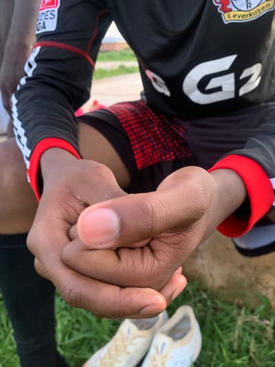 The image shows a close-up view of a person's hands, one fist clenched and the other partially open, against a backdrop of green grass and some dirt. The person is wearing a black sports jersey with red accents on the sleeves and collar. Visible text on the jersey includes "Leverkusen" and what appears to be the number "23" in large white font. There is also a red and black checkered pattern on the fabric. The lighting suggests it could be daytime. The setting appears to be outdoors, possibly a sports field, given the attire and the grass. The focus is on the hands and the texture of the skin, with some of the sporting apparel visible. The crops provide closer views of the jersey details and the hand's knuckles and fingernails.