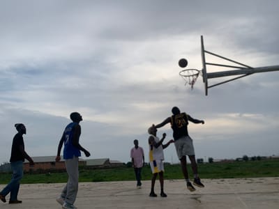 The image depicts a group of young men playing basketball on an outdoor court. The sky is overcast, suggesting a cloudy day. The scene appears to be in an open area, with buildings visible in the background and some sparse vegetation. The court surface is concrete.
Five individuals are visible. In the foreground on the left, a man in a black t-shirt and blue jeans walks towards the left side of the frame, looking to his left. Next to him and slightly behind, a man in a blue basketball jersey with the number "7" on the back, and grey pants, walks towards the right. In the center, two men are engaged in the game. One, wearing a white jersey with yellow trim and yellow shorts, is in the air, reaching for the ball. Another man, wearing a black jersey with "24" on the back and grey shorts, is also in the air, possibly attempting to block the shot or jump for a rebound. Behind them, a man in a pink shirt walks away from the camera. A basketball is visible in the air, near the hoop.
The overall mood seems casual and active, indicative of a friendly game. The time of day is likely late afternoon or early evening, given the diffused light and the cloud cover. The presence of buildings and the open, undeveloped landscape suggest a location in a less urbanized area. There is visible text on the blue jersey, partially obscured but appears to say "GARRETT" above the "7". The text on the black jersey reads "24".
