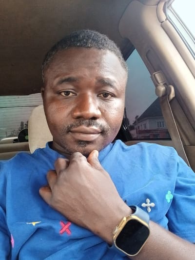 A dark-skinned man is in a car. He is wearing a blue shirt with embroidered designs and a smart watch with a gold case and grey strap. He is sitting in the driver's seat, and his right hand is near his neck. A seatbelt is visible. Outside the car window, buildings are visible. This is likely in Dushepe, Nigeria.