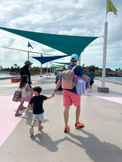 The image depicts a family of four walking along a paved pier on "Perfect Day at CocoCay," Berry Islands, Bahamas, under a bright, partly cloudy sky. The pier, predominantly light grey with a narrow pink stripe on its left side, is shaded by multiple triangular tensile fabric structures in teal, dark blue, yellow, and green.

From left to right, walking away from the camera, are: a woman with dark hair wearing a dark top, purple baseball cap, and light sandals, carrying a patterned tote bag; a young girl in a light floral top, pink shorts, and sandals; a young boy in a black t-shirt, patterned swim shorts, sandals, and a neck lanyard, holding the hand of an adult male; and the adult male, seen from the back, wearing a light blue baseball cap with an anchor logo, a dark blue t-shirt, coral shorts, and bright orange and black athletic shoes. He carries a tan backpack and a light blue rolled towel over his left arm.

White poles supporting the shade structures also display flags, including a dark flag (possibly a pirate flag) on the left and a light green flag on the right. Calm, light blue water is visible on both sides of the pier. In the background on the left, "PERFECT DAY" signage is visible among palm trees and distant land. Further down the pier on the right, a staff member in uniform stands, facing the approaching family. Distinct shadows of the individuals and structures are cast upon the pier's surface.