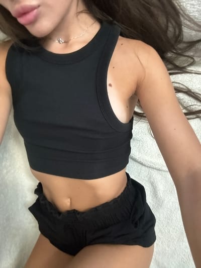 This is a close-up, overhead shot of a person lying on a textured, white surface. The person has long, dark hair that extends across the frame. They are wearing a black tank top and black shorts. A delicate silver necklace with a small pendant is visible around their neck. The lighting is soft, suggesting an indoor setting. The person's skin tone is fair with visible moles on their shoulder and arm. The overall mood is relaxed and casual. There is no visible text in the image.