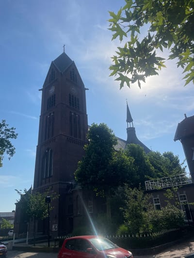 This media file captures an outdoor scene under a bright, sunny sky, with the sun's rays casting a lens flare. The primary subject is a tall, dark brick church tower with a pointed roof and a cross at its apex. The tower features multiple arched windows and at least two visible clock faces. Surrounding the tower are lush green trees, with some branches and leaves of a deciduous tree in the foreground, partially obscuring the view. To the right, a portion of a red car is visible, parked alongside a sidewalk and a decorative fence. In the background, part of a building with windows and a pitched roof can be seen. The sky is a clear blue with some wispy white clouds. The overall impression is of a tranquil, daytime scene in a European urban or semi-urban setting, possibly Venlo, Netherlands, given the context. The orientation of the image is such that the church tower appears to be leaning towards the viewer. The image is captured from a low angle, looking up towards the sky and the top of the church.