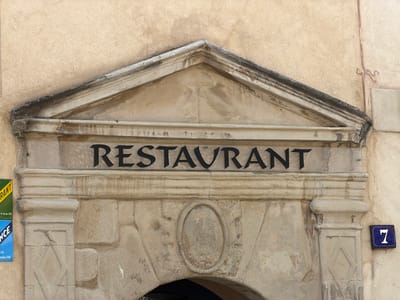 The media file shows a close-up, eye-level exterior view of an old stone building with a sign that reads "RESTAURANT". The sign is made of black, stylized lettering on a transparent background, mounted above an arched entryway. The stone facade of the building is weathered and textured, with a triangular pediment above the sign. Below the sign, there is a decorative carved oval in the stone arch. To the left of the entryway, there are two colorful signs, one yellow with green lettering and the other blue with white lettering. To the right of the entryway, a small blue sign with the number "7" in white is affixed to the wall. The building appears to be located in a historical district, possibly in Mercurol-Veaunes, France, given the context. The overall impression is one of history and tradition. There is no indication of people or specific weather conditions.