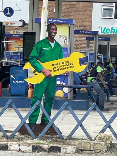 The image shows a life-sized cardboard cutout of a smiling Black man in a green uniform holding a large yellow wrench. The cutout is standing in front of a gas station in Ilado, Nigeria. The gas station has multiple fuel pumps and signage for "ENVO" and "SHELL HELIX." In the background, several other people, some in similar uniforms, can be seen near the pumps. A dark-colored car is partially visible on the left side of the image. The scene appears to be outdoors, under an overhang, with brick buildings visible in the background. The foreground shows a blue metal barrier and some debris on the ground. The cutout has text on the wrench that reads "Drive in to get your car serviced in 25 mins or less."
