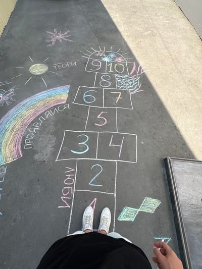 The image shows a hopscotch court drawn with chalk on an asphalt surface. The hopscotch grid is numbered from 1 to 10, with some of the squares decorated with drawings of flowers and plants. There are also drawings of a sun, a rainbow, and some abstract shapes. Text written in Cyrillic letters is visible, including "ТВОРИ" (create) and "ЛЮБИ" (love), and part of "ПРАВДА" (truth). The scene appears to be outdoors, possibly on a sunny day, given the shadows cast on the ground. A person's legs and feet, wearing white sneakers and black clothing, are visible in the lower right corner, suggesting they are either playing hopscotch or observing it. The overall mood seems playful and creative. The location is identified as Simferopol, Ukraine.