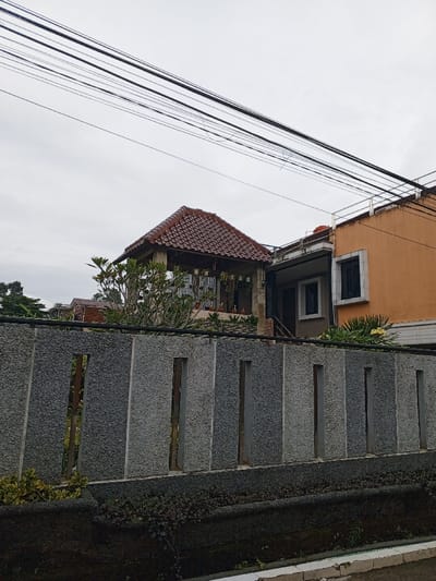 A residential scene with a gray stone fence in the foreground. Behind it, there's a house with a distinctive red-tiled gazebo-like structure and an adjacent orange building. The sky is overcast, and a network of overhead wires cuts across the top portion of the frame. Some greenery is also visible on and around the fence.