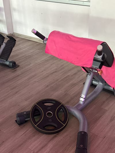 The image shows a close-up, slightly angled view of a piece of gym equipment, likely a weight-lifting machine. A bright pink towel is draped over a portion of the machine, covering part of its metallic structure. A black and yellow weight plate, marked with "15," is attached to the machine. The equipment rests on a wooden floor with a visible grain pattern. In the lower left corner of the image, part of another piece of gym equipment, possibly a leg press or similar machine with black footpads, is visible. The background appears to be a white wall, with a hint of a window or partition on the left. The lighting suggests an indoor setting, likely a gym. The overall scene is static, with no people visible, and the focus is on the gym equipment and its accessories. The text "MATRIX" is visible on the metallic frame of the machine. The context provided states the location is Rathenow, Germany, but there are no explicit visual cues in the image to confirm this.
