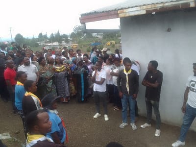 A diverse crowd gathers outside a plain, rectangular building with a corrugated roof. People wear a mix of casual and formal attire. Several individuals stand near the building's wall, some wearing yellow and black scarves. In the background, houses are visible, and a yellow vehicle is parked on a distant road. The atmosphere appears to be one of anticipation or waiting.