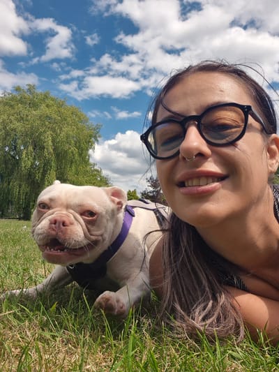 This is a selfie taken outdoors in Bridlington, United Kingdom, on a sunny day with a blue sky and scattered clouds. A woman with long brown hair, wearing black-rimmed glasses and a nose ring, is smiling at the camera. Beside her, a white French bulldog with a purple harness is lying on the grass. A large tree with green foliage is visible in the background. The setting appears to be a park or grassy area within the city.