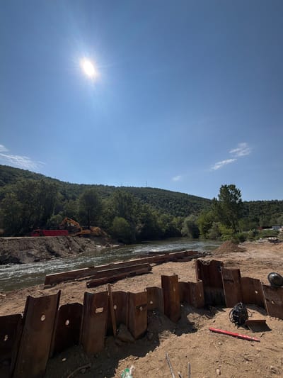 The image depicts a construction site by a river in Kresna, Bulgaria. In the foreground, a wall of metal sheeting, likely sheet piling, is partially driven into the earth. Various construction materials, including rebar, a level, and a welding machine, are scattered on the sandy ground. Further back, a river flows, with construction equipment including a red truck and a yellow excavator visible on the embankment. The background is dominated by a lush, green, tree-covered hillside under a bright blue sky with a prominent sun. The scene suggests ongoing riverbank reinforcement or construction work.