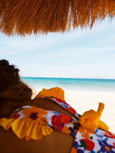 The image shows a person in a floral bikini lying on a white sandy beach under a thatched straw parasol, looking out at the sea in Ras Abu Soma, Egypt. The sea is light blue and there is a light blue sky with some wispy clouds.