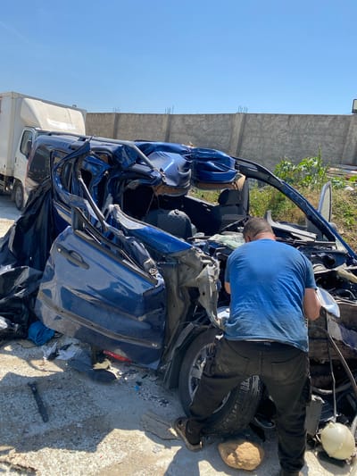 The image shows the aftermath of a severe car accident. The location is identified as Baraki, Algeria. A blue vehicle has sustained extensive damage, with crumpled doors, a crushed hood, and a distorted frame. A man in a blue shirt and dark pants is seen leaning over the vehicle's engine compartment, seemingly examining the wreckage. The car rests on a concrete surface with debris scattered around it. A white truck is partially visible in the background, suggesting the scene might be a junkyard or salvage yard. A concrete wall stands in the background, possibly indicating an urban environment. The focus is on the wrecked vehicle and the man's interaction with it.