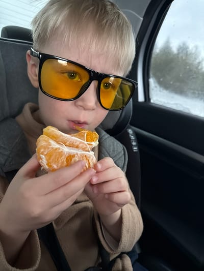 A blonde-haired child wearing large, black-rimmed sunglasses with yellow lenses is reclining in a car seat and eating a peeled mandarin orange. The child is wearing a beige or light brown sweater. The background includes a window with rain or moisture on the glass and part of the car's interior. The child's car seat is gray and appears to be secured with straps. The photo suggests the scene is inside a moving vehicle, potentially in transit. The information regarding the location in Vakhrino, Russia, cannot be verified from the visual content of the image.