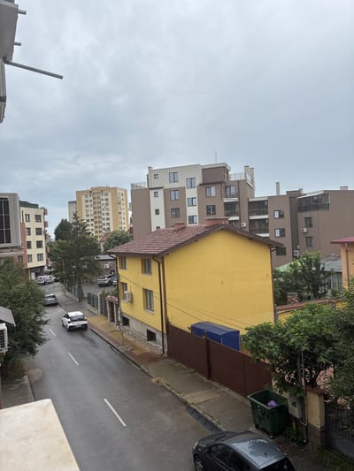 The image depicts a view of a street in Kazanlak, Bulgaria, on an overcast day. The street is wet, suggesting recent rain. Several vehicles are visible: a white hatchback parked on the street, a darker colored hatchback parked closer to the foreground, and a silver SUV parked further up the street. Buildings of varying heights line the street. A prominent yellow two-story house with a red tile roof is in the foreground. Behind it, taller apartment buildings, some with light brown and beige facades, rise up. Trees and greenery are interspersed throughout the scene. A large green trash bin and a blue tarp are visible near a brown fence. The overall impression is of a residential urban environment.