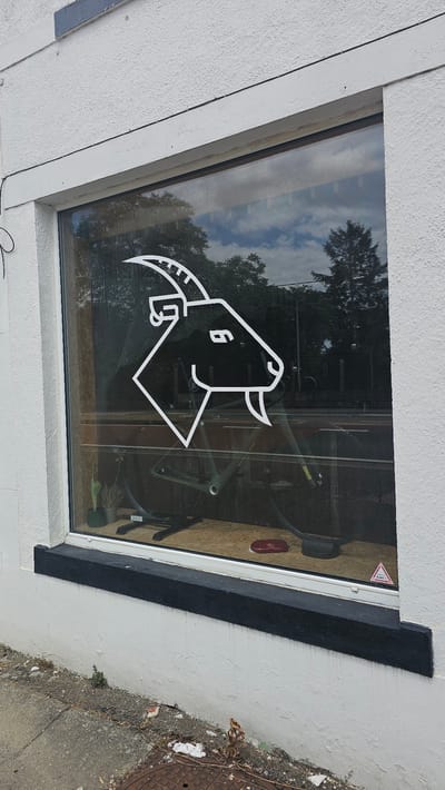 The image shows a storefront window with a white outline logo of a goat's head. The logo is superimposed on the reflection of trees and sky in the window. Inside the store, a bicycle is visible, mounted on a stand. A small potted cactus sits to the left of the bicycle. The building appears to be made of textured white stucco, with a black painted windowsill. The exterior ground is a mix of concrete and gravel, with some debris and vegetation. A manhole cover is visible in the lower right corner. The setting is likely a street in a town or city, though no specific street signs or identifying landmarks are present in the image that would confirm its location in Boussu-lez-Walcourt, Belgium.