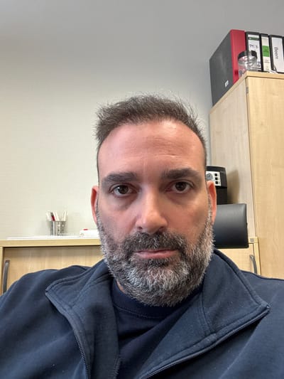 A close-up headshot of a middle-aged man with graying hair and a beard is visible. He is wearing a dark blue zippered jacket. Behind him, there is a light-colored cabinet with several binders and a black telephone on top. The wall is light gray. There is a metal bracket with papers attached to the wall. There is not enough information to accurately determine the location to the city of Grevenmacher, Luxembourg.