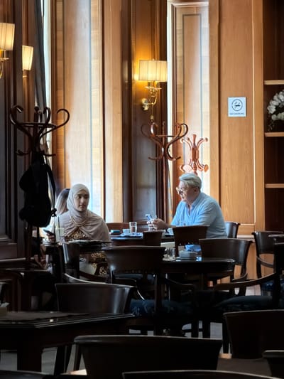 The image depicts the interior of a restaurant or cafe, likely in Sarajevo, Bosnia and Herzegovina, characterized by its warm, wood-paneled walls and classical decor. Two individuals are seated at a table in the foreground. On the left, a woman wearing a hijab and a pearl necklace is looking down at her phone. On the right, a man with glasses and a light blue shirt is also looking at his phone. The table is set with drinks and possibly food items. In the background, there are decorative coat racks, ornate lamps, and a "No Smoking" sign on the wall, suggesting a non-smoking establishment. The lighting is soft, creating a cozy ambiance. The overall scene captures a moment of quiet interaction or individual focus within a public setting.