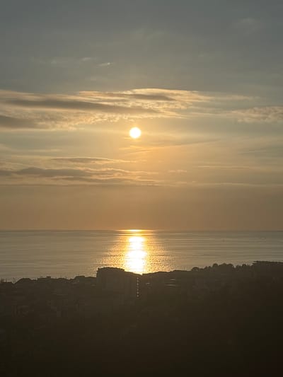 The image depicts a serene coastal sunset scene in Adlerskiy, Russia. The sun, a bright orb, hangs low in the sky, casting a warm, golden glow that reflects off the calm waters of the sea. Wispy clouds streak across the sky, illuminated by the sun's rays, creating a dramatic and ethereal atmosphere.

On the right side of the image, a dark silhouette of the coastline rises from the water. Buildings and structures are faintly visible against the darkening sky, suggesting a town or city nestled along the shore. The overall mood of the image is peaceful and tranquil, evoking a sense of awe at the beauty of nature. The time of day is clearly sunset, and the weather appears clear with some light cloud cover. The location is identifiable as Adlerskiy, Russia, likely a coastal area with buildings along the shore. No text is visible in the image.