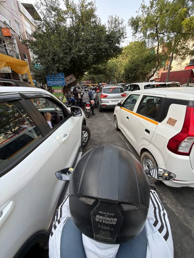 The image depicts a traffic jam on a street in Delhi, India. In the foreground, the back of a person wearing a black helmet and a white and black striped shirt is visible, suggesting the photo is taken from a motorcycle. Several white cars are lined up, with a white SUV on the left and a white hatchback on the right. A few motorcycles and scooters are also present in the traffic. In the background, trees with dense foliage line the street, and buildings with a mix of brick and painted facades can be seen. Signs are visible on some of the buildings, and a yellow awning is draped over one. The sky is overcast. The presence of vehicles with Indian license plates and the general streetscape are consistent with a location in Delhi.