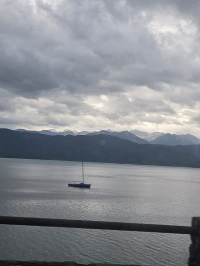 A sailboat is anchored on a calm lake in front of a tree-covered mountain range under a cloudy sky. The lake is dark blue and reflects the overcast sky. The mountains in the background are dark green and covered in trees. The sky is filled with gray clouds. A wooden railing runs along the bottom of the image. The scene is a serene and peaceful landscape.