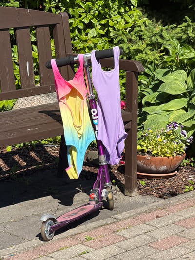 In Baloži, Latvia, a purple kick scooter is parked on a paved walkway next to a wooden park bench. Two swimsuits are hung over the handlebars of the scooter: one with a pink, yellow, and blue gradient pattern, and another solid lavender swimsuit. Behind the bench, lush green foliage creates a natural backdrop, with a terracotta pot filled with yellow and purple flowers to the right. The scene is bathed in bright sunlight, casting shadows on the walkway and the ground.