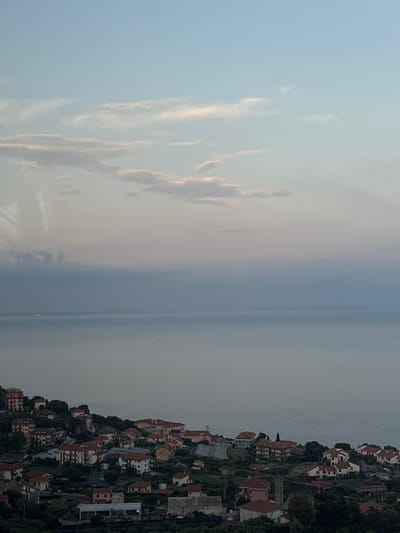 A coastal view shows a town nestled on a hillside, with closely packed buildings and terracotta roofs. The sea stretches to the horizon under a cloudy sky. Light filters through thin clouds, creating soft hues in the sky. The atmosphere is calm and slightly hazy, suggesting a tranquil coastal environment.