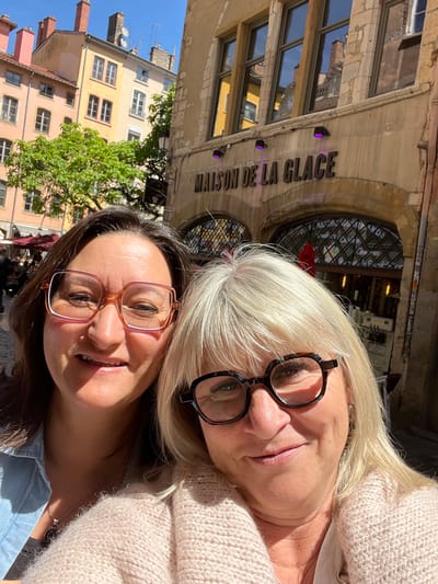 Two women are posing for a selfie. The woman at the bottom of the frame has brown hair and is wearing pink-framed glasses and a blue shirt. The woman at the top of the frame has blonde hair and is wearing black-framed glasses and a light pink sweater. Behind them is a building with the sign "MAISON DE LA GLACE". The building appears to be in Lyon, France, given the architectural style. Other buildings and a tree are visible in the background, along with a blue sky.