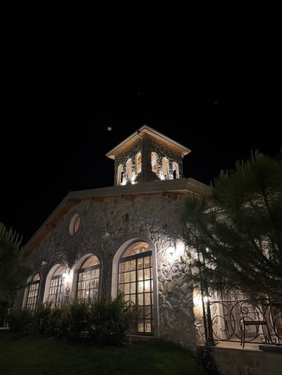 The image shows a stone building at night, illuminated by external lights. The building has a rustic appearance, with a tower topped by a pitched roof. Large arched windows with multiple panes are visible on the facade, and a circular window is present on the upper level. A wrought-iron balcony with a chair is visible on the right side of the building, partially obscured by a pine tree. The ground in front of the building is covered with grass. The location is identified as the town of Kocherinovo, Bulgaria, specifically the St. George the Conqueror winery.