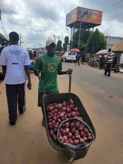 In Egbagu, Nigeria, a man pushes a wheelbarrow overflowing with red onions down a dusty road. He wears a green t-shirt and a beige cap. Beside him, another man in a white t-shirt with "BALENG IAGA FASHION MEN'S WEAR" printed on it walks ahead. The background shows a busy street scene with cars, motorcycles, and pedestrians. A large billboard advertising "Lovebite Family Ahead" is visible in the distance. The sky is cloudy.