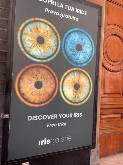 The image shows a rectangular sign displayed on the street level, likely in Bologna, Italy, based on the Italian text. The sign advertises "Irisgalerie" and offers a "Free trial" or "Prova gratuita" to "Discover your iris" or "Scopri la tua iride." The sign displays four images of irises, each ringed with a colored circle. The irises are colored blue, yellow, a mix of yellow and blue, and brown. The sign is black with white text. The sign is situated under a wooden door frame.