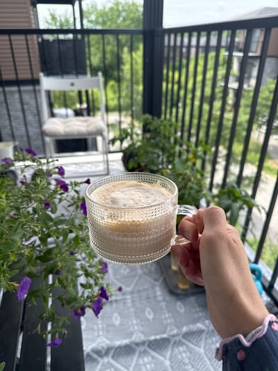 A hand holds a glass mug of coffee on a balcony in Gatineau, Canada. The mug has a textured glass design. In the foreground, purple flowers and green foliage are visible. In the background, a white chair with a cushion is present, along with a black railing and trees. The sky is overcast.