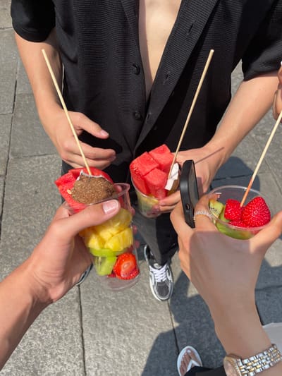 Four people sit on the ground outside in Verona, Italy. They each hold a clear plastic cup containing fruit on a stick. The fruits include watermelon, strawberry, kiwi, pineapple, and coconut. One person is wearing a black shirt, one person is wearing black and white sneakers, and another person is wearing a watch. The people's legs are visible. One person is holding a black phone. The ground is a grey paved surface.