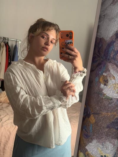 A young woman is captured in a mirror selfie, holding an orange smartphone to take the photo. Her gaze is directed towards the camera, and she wears a neutral expression. She has light brown hair styled with bangs and is dressed in a white, textured blouse featuring ruffled, voluminous sleeves and delicate lace detailing at the cuffs and collar. Rings are visible on her ring fingers, and an Apple Watch with a patterned band peeks out from beneath her left sleeve.
The smartphone case has a sticker on its back that partially displays text, appearing to be "den" or "uen," accompanied by smaller letters "RM" and a decorative swirl. The background of the selfie reflects an indoor setting, likely a private room such as a bedroom or dressing area. To the left, a clothing rack with various garments is visible. Below the woman, light blue fabric, possibly a skirt or pants, is seen, alongside light-colored bedding. Above her head, a blurred reflection of a patterned fabric or object is visible, consistent with a reflection in a mirror. The image was originally rotated 90 degrees clockwise. The setting for this image is Большая Шильна, Russia.