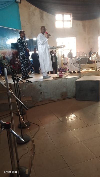 The scene depicts an indoor public gathering in Ube, Nigeria. A middle-aged male, wearing a white traditional tunic and glasses, stands on a raised platform addressing an unseen audience. He holds a red-capped microphone in his right hand, positioned behind a clear lectern. To his immediate left, a male in a camouflage-patterned uniform stands at attention. Several other individuals are visible in the background and to the right, some seated on white and gold upholstered chairs/sofas, others standing. A prominent multi-pane window in the rear wall provides strong natural backlighting. The floor throughout the venue and on the platform is covered with light-colored square tiles. In the foreground, multiple microphone stands are positioned, with cables running across the floor. A large, dark grey speaker box rests on the floor to the right of the platform. Decorations include blue and black fabric drapes overhead, a large blue banner on the left, and a small arrangement of purple and pink artificial flowers on the platform. A caged standing fan is also visible on the platform to the right. The venue walls appear to be light-colored, showing some signs of age or wear.