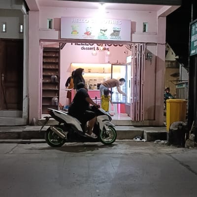 The scene takes place in front of a shop called "Hello Smoothies & Dessert & Cake" in Kaliwungu, Indonesia (latitude: -6.963, longitude: 110.251). A man is seated on a white motorcycle with a green rimmed wheel. He is wearing a black shirt and black pants. A woman in a white headscarf stands near the entrance to the shop. She is wearing a blue shirt and black pants. A man in a green shirt is standing near the window of the shop. He is facing away from the camera and looking at something inside the shop. A small child in a yellow shirt is standing next to him. There is a yellow trashcan with a lid on the right side of the image. The sidewalk is made of light-colored concrete, and the ground is paved with asphalt. The store has a pink facade and a glass front. A sign above the store reads "HELLO Smoothies". The storefront is decorated with a menu for smoothies and desserts. The environment is dimly lit and the lighting appears to be from streetlights.