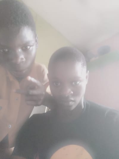 Two young Black males, appearing to be teenagers, are captured in a selfie-style photograph. The individual in the foreground, on the right side of the frame, is wearing a dark t-shirt and looking directly at the camera. The second individual is positioned behind and to the left of the first, wearing a tan collared shirt. This person's left hand is extended, with a finger pointing towards the viewer, seemingly interacting with the person taking the photo. The background appears to be an indoor domestic setting, with plain walls. The lighting is bright and somewhat diffuse, creating a slightly overexposed and hazy effect across the image. There are no identifiable landmarks or elements in the image that would allow for the specific determination of a location within Shangisha, Nigeria. The image does not provide sufficient detail to offer any further forensic analysis or specific contextual information about the scene or the individuals.