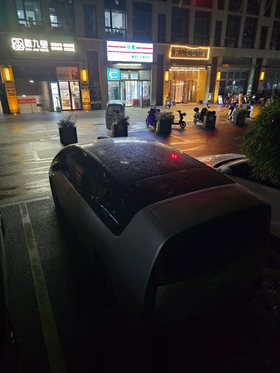 The image depicts a nighttime scene on a wet street in the Xiaoshan District of China. The setting appears to be an urban area with several businesses lining the street. Visible signs include "風離九堂 ZHI JIU TANG", "东方之新", "开元名城1幢", and "城市金烤便利店". A license plate on a vehicle reads "池A-C58758". Several parked motorcycles and scooters are scattered along the sidewalk, suggesting a mode of transportation common in the area. The ground is damp, indicating recent rain. The lighting suggests it is late at night, with artificial lights from the buildings illuminating the street. The overall mood is quiet and slightly reflective, amplified by the wet conditions.
