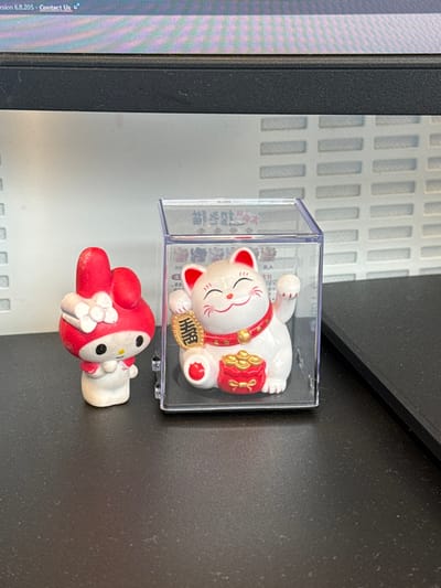 In Sint-Niklaas, Belgium, a forensic examination reveals two figurines resting on a dark, textured surface showing dust particles. To the left is a small, off-white figurine with a red hood and ears, consistent with the character My Melody, featuring a white bow on its head. Its surface exhibits minor wear and dirt. To the right, a white Maneki-neko figurine is enclosed within a clear plastic display case. The Maneki-neko has a red collar with a golden bell, a raised left paw holding a golden oval coin with an East Asian character, and a red sack overflowing with golden coins at its base. Printed text, likely in an East Asian language, is visible within the display case. The background consists of a light-colored wall with horizontal slotted panels. Above the panels, the lower portion of a computer monitor screen is visible, displaying digital text including "sion 6.8.205 - Contact Us".